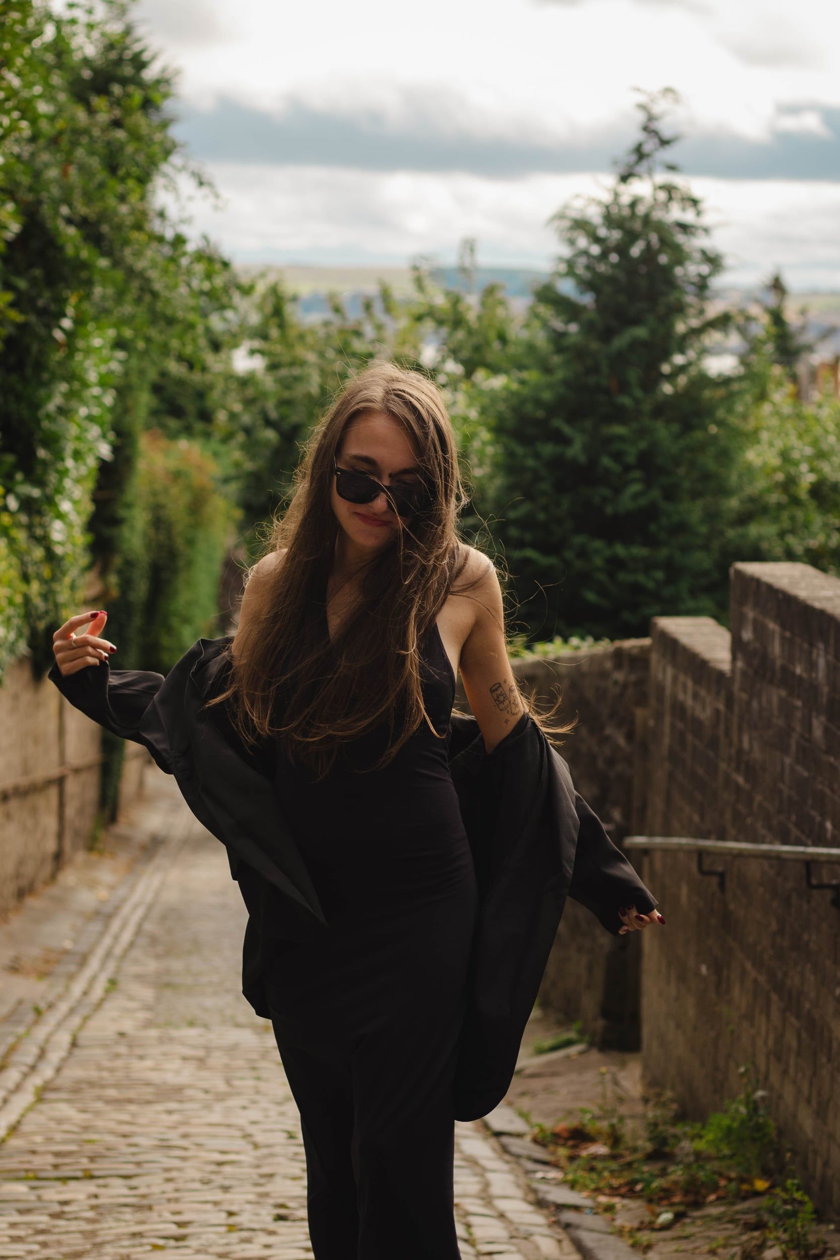 Woman in a black dress and sunglasses on a cobbled lane, jacket slipping off her shoulders