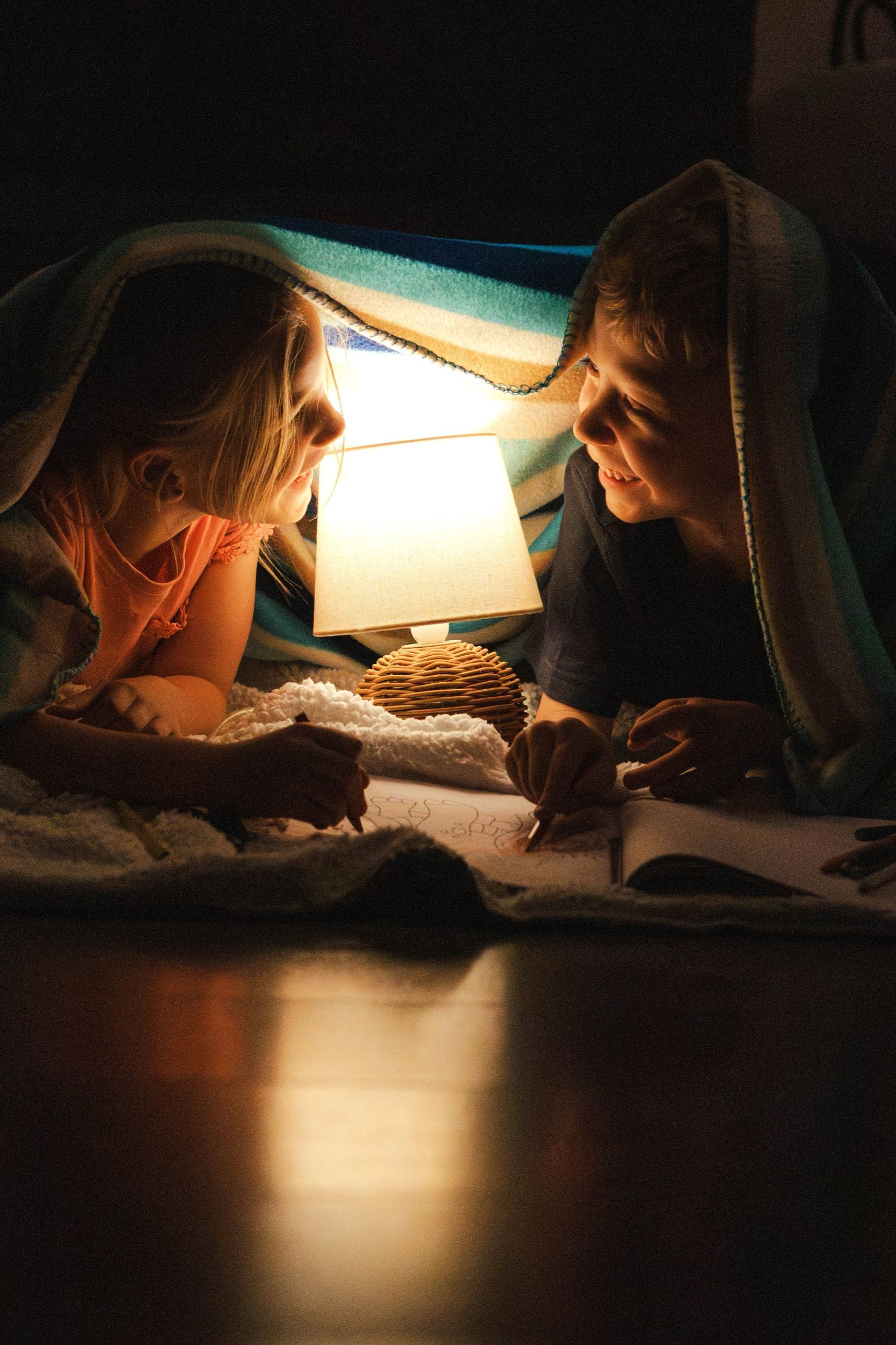 Two children drawing under a blanket tent, lit by a small lamp