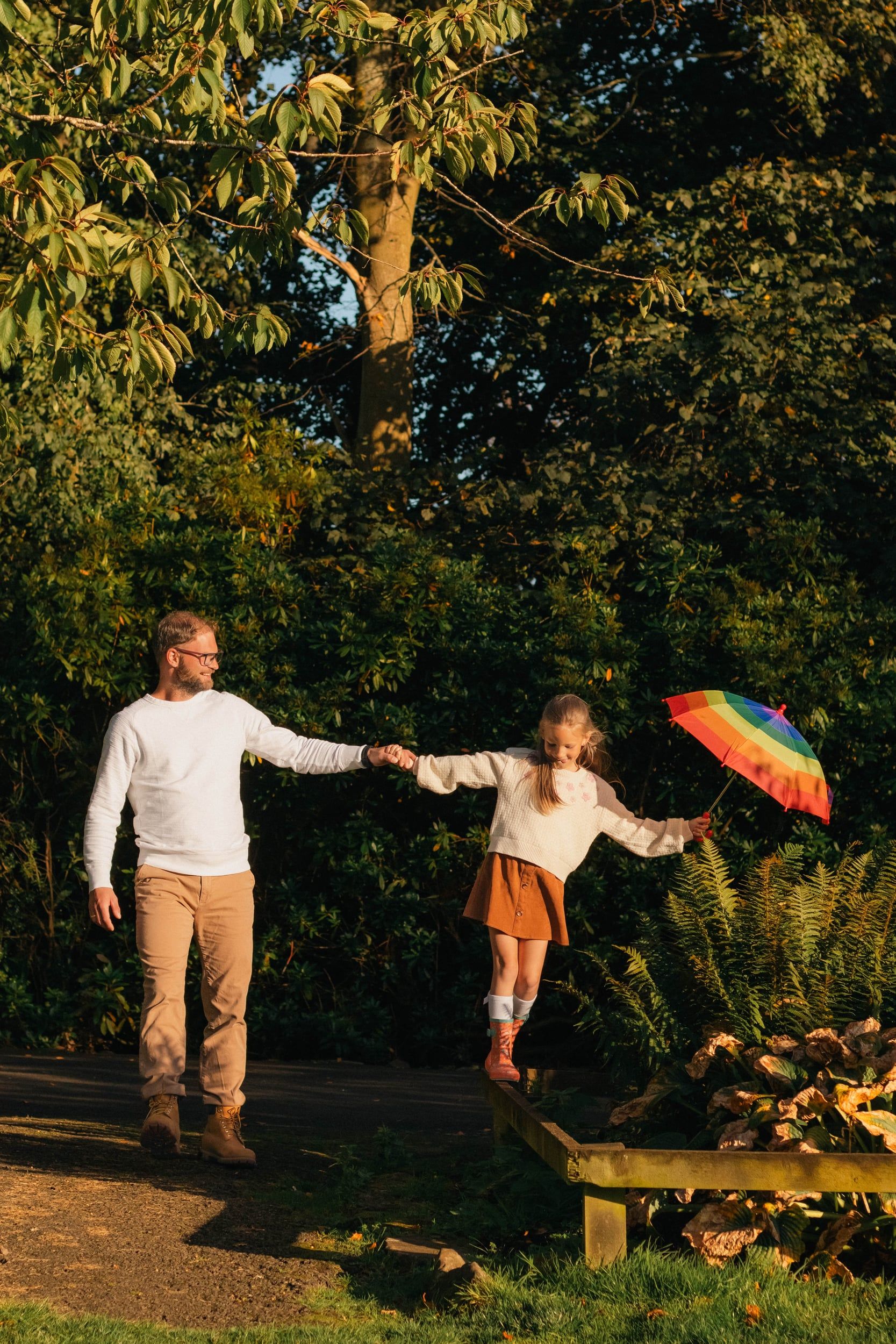 Father holding his daughter’s hand as she balances on a log with a rainbow umbrella