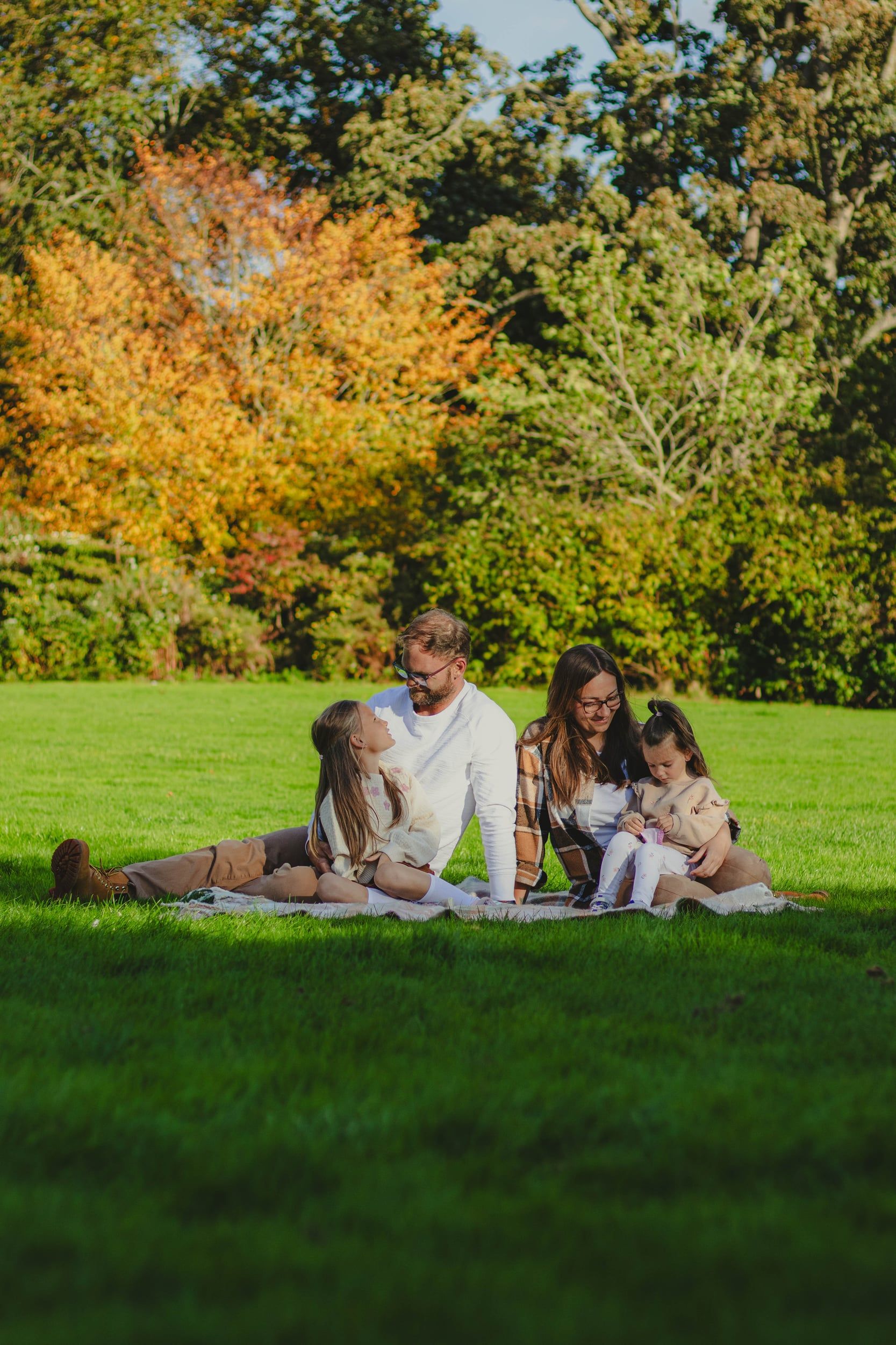 Family of four sitting on a blanket on the grass, autumn trees behind