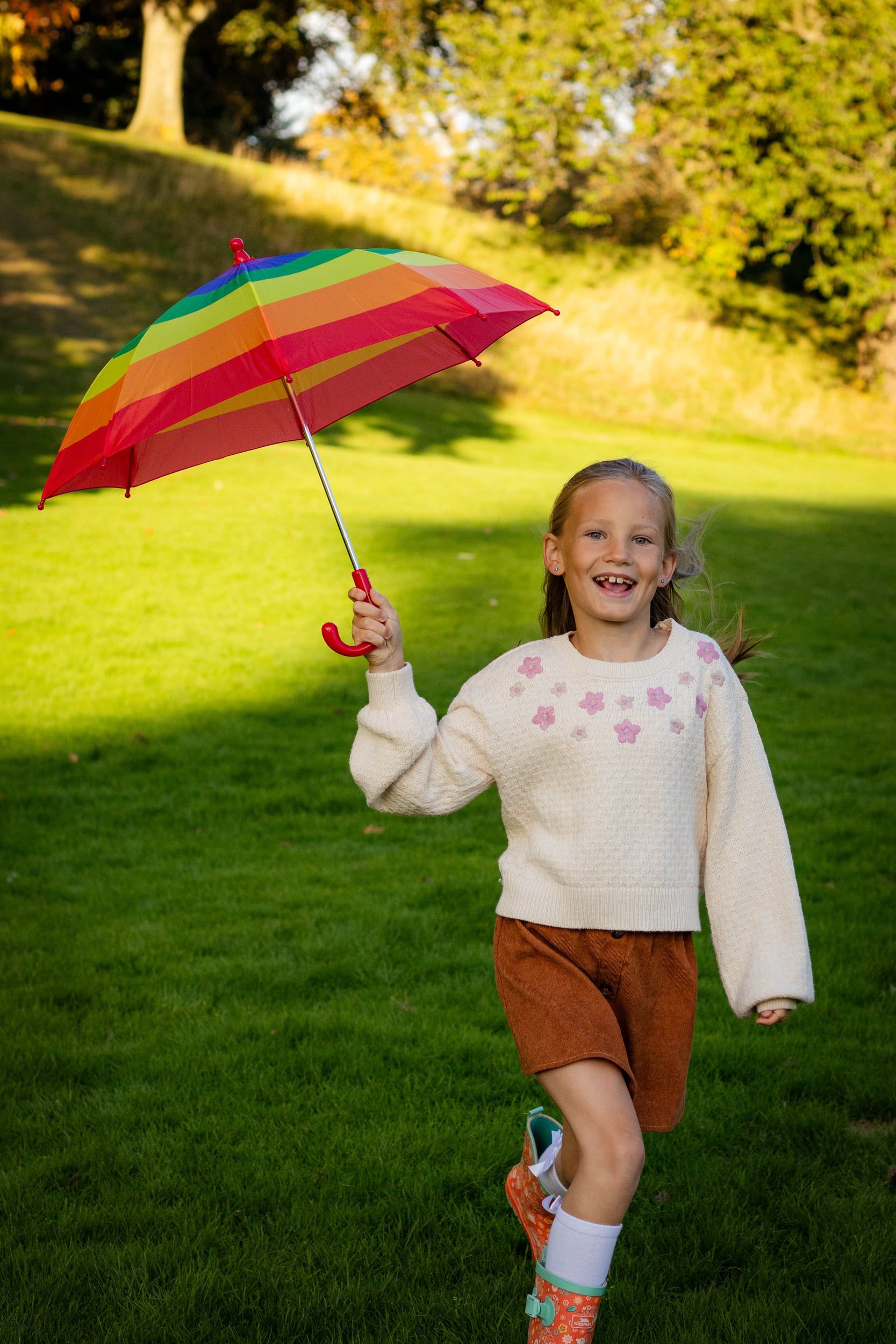 Girl running across sunny grass holding a rainbow umbrella