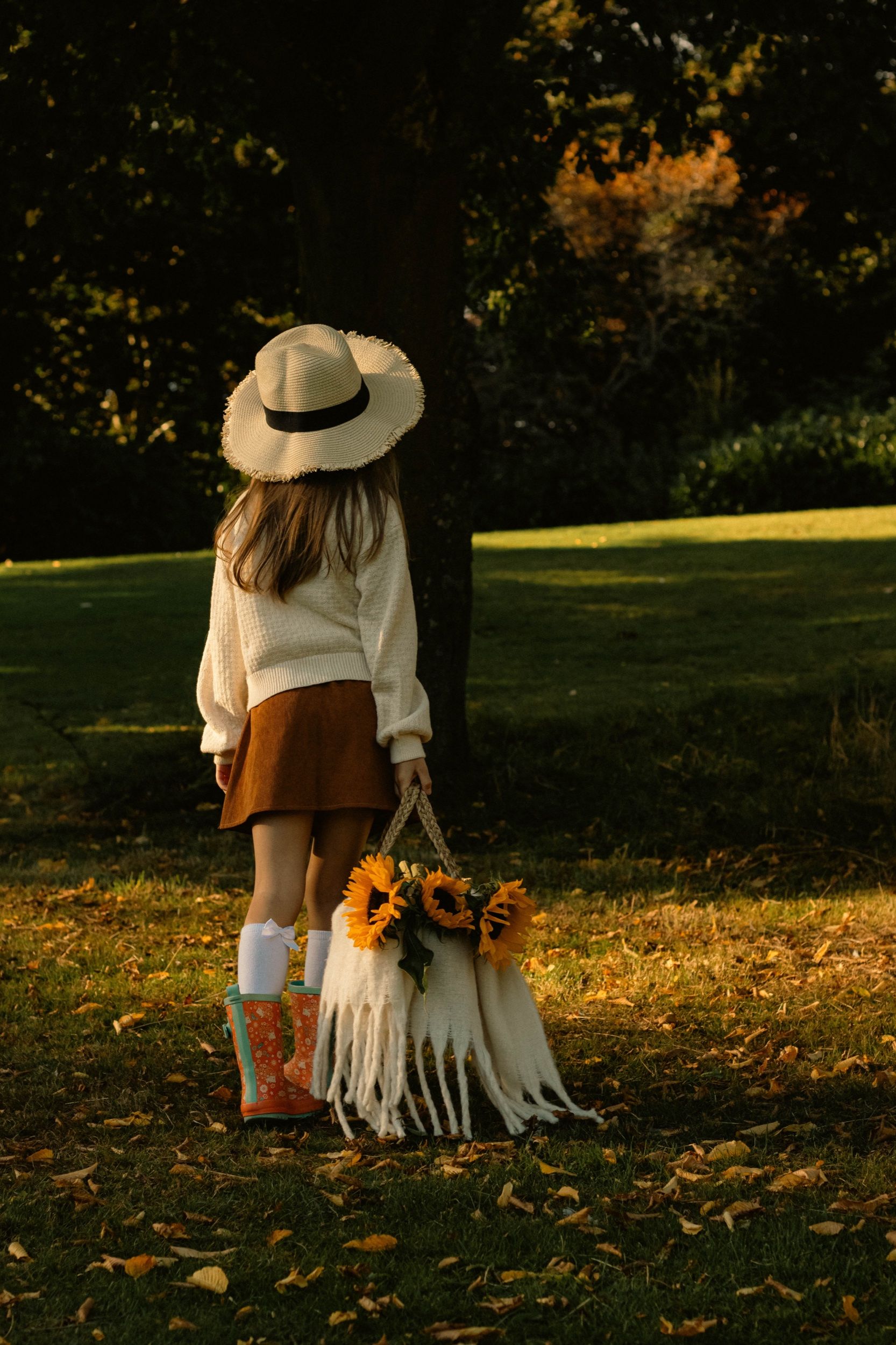 Girl in a straw hat and wellies carrying a basket of sunflowers across the grass